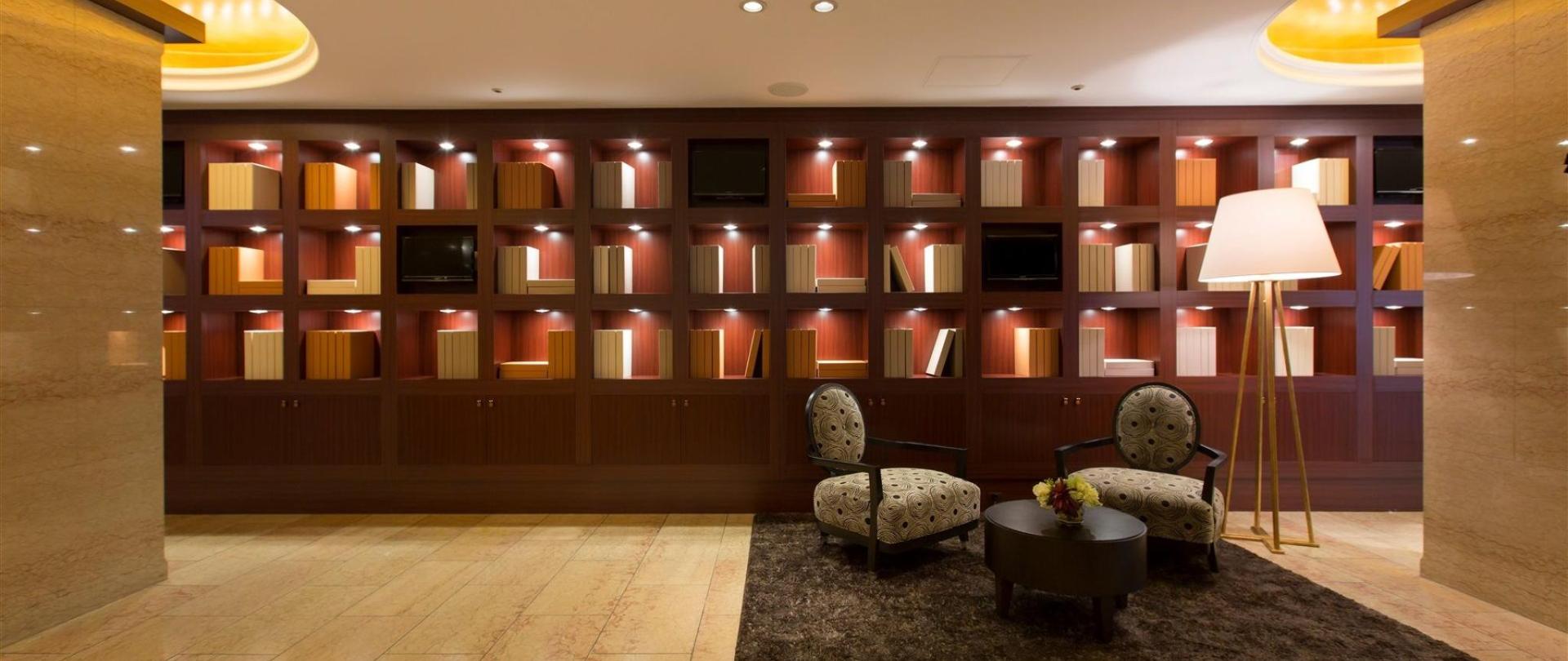Interior lobby with a large wall of lit display shelving resembling book spines, two patterned armchairs, and a floor lamp