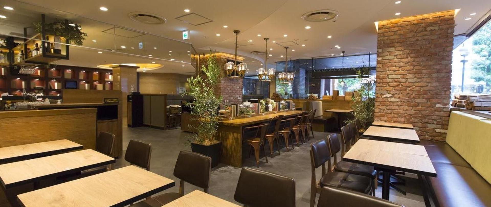 Modern cafe or restaurant interior with wooden tables, dark chairs, exposed brick columns, potted plants, and a long communal counter