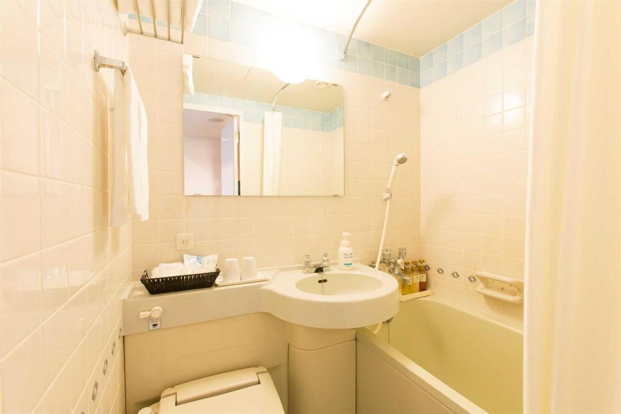 A compact hotel bathroom with pale tiled walls, a sink, mirror, toilet, and a bathtub with handheld showerhead
